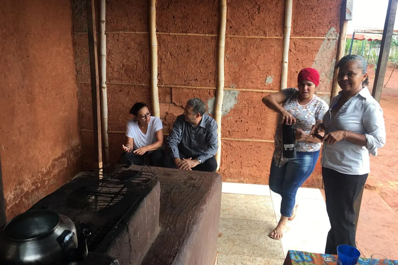Quatro adultos conversam dentro de uma construção de barro com fogão a lenha.