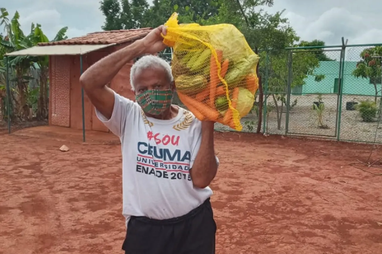 Senhor de máscara carrega no ombro uma sacola de laranjas e cenouras em um pátio de terra vermelha.