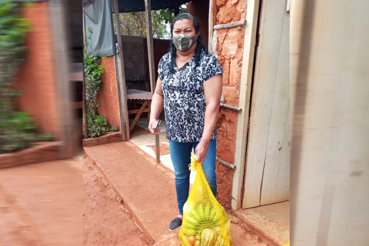 Mulher de máscara segura uma sacola amarela de alimentos diante de uma construção de barro.
