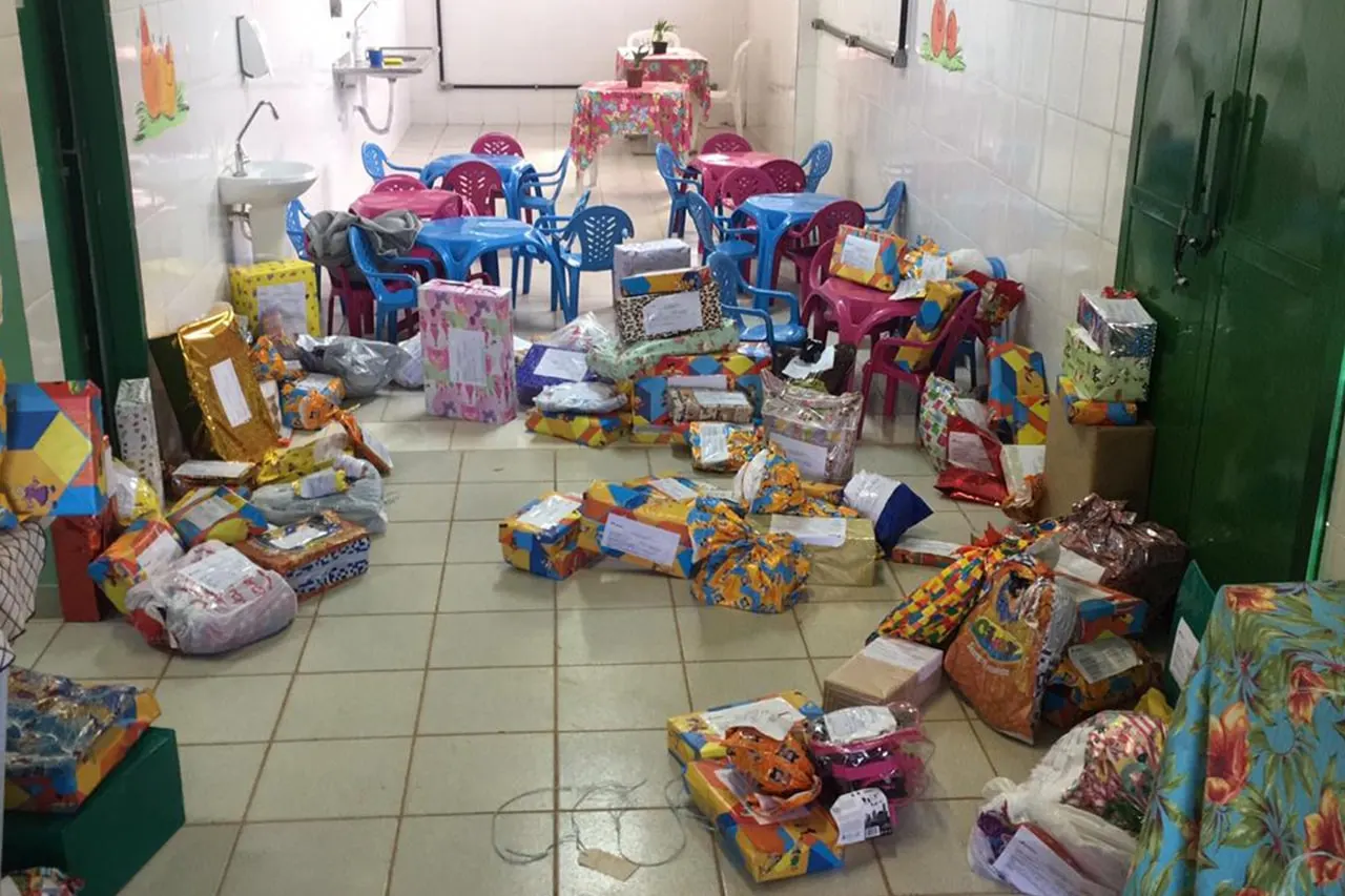 Sala com mesas pequenas e muitos pacotes, brinquedos e cestas de doações no chão.