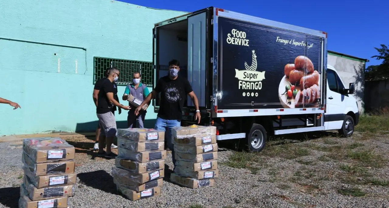 Caminhão de entregas ao lado de pilhas de caixas de alimentos, com homens de máscara descarregando.