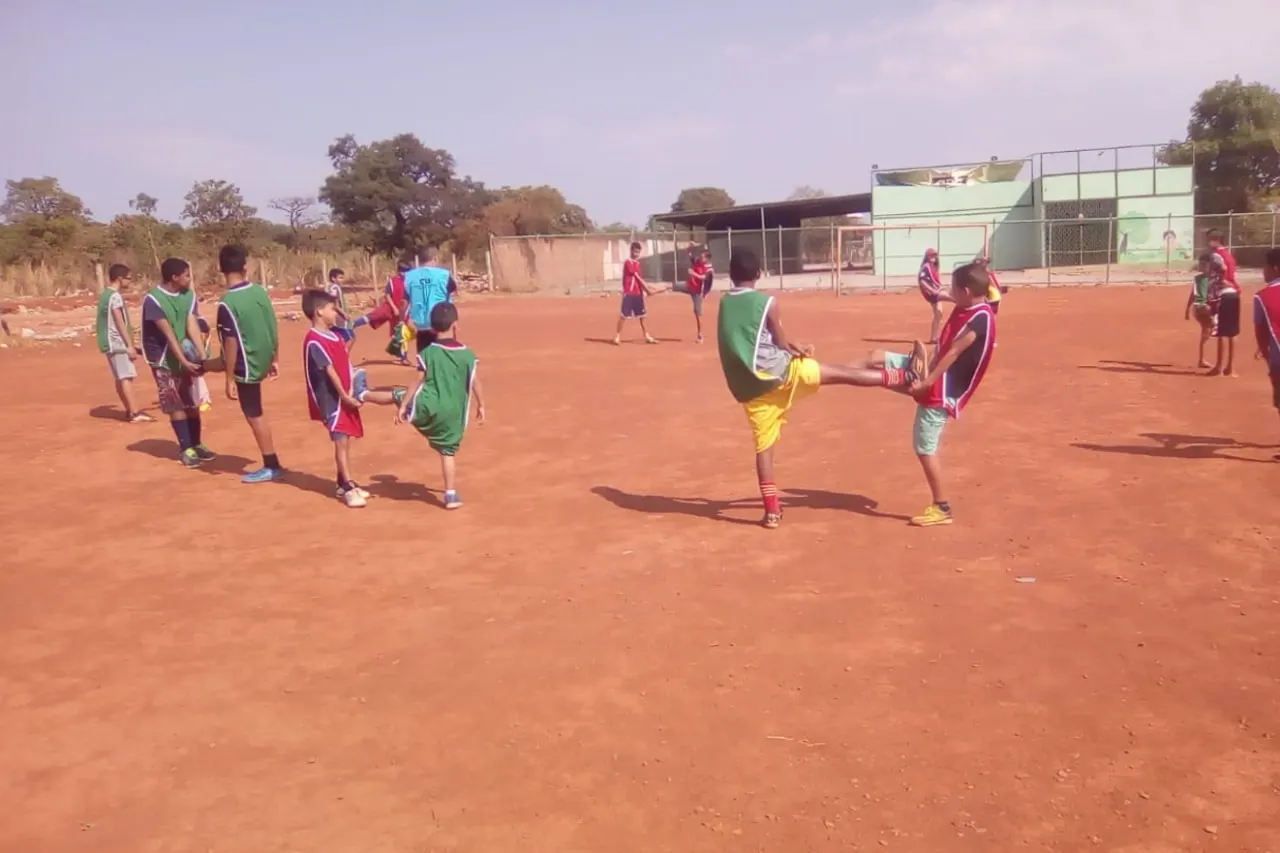 Crianças correm atrás da bola em um campo de terra vermelha, vistas de longe.
