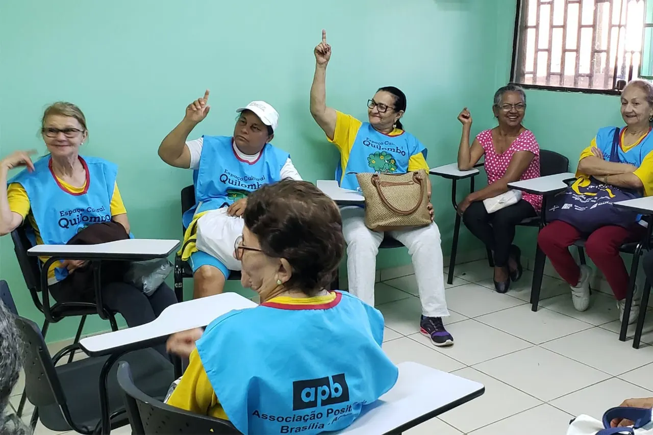 Idosos de colete azul em uma sala de aula de paredes verdes, alguns com a mão levantada.