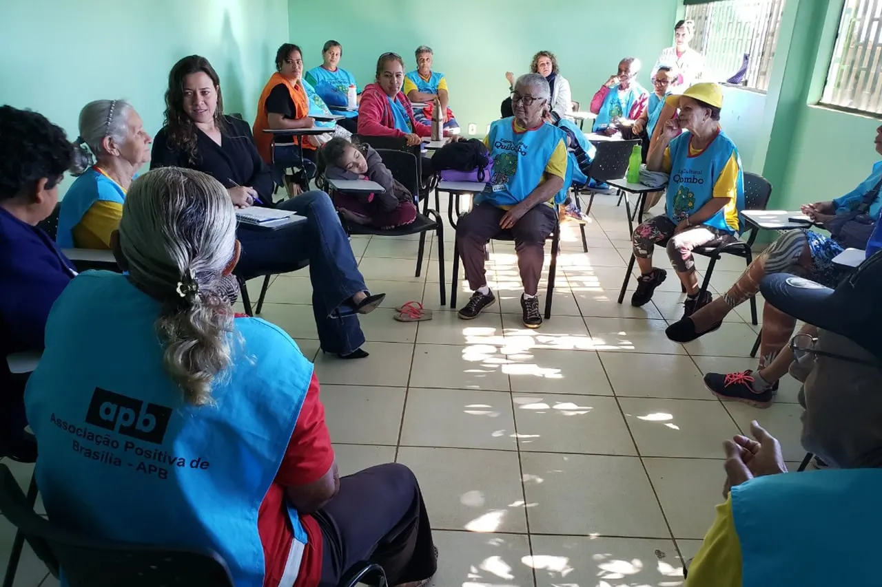 Idosos de colete azul sentados em círculo em uma sala de paredes verdes durante uma atividade.