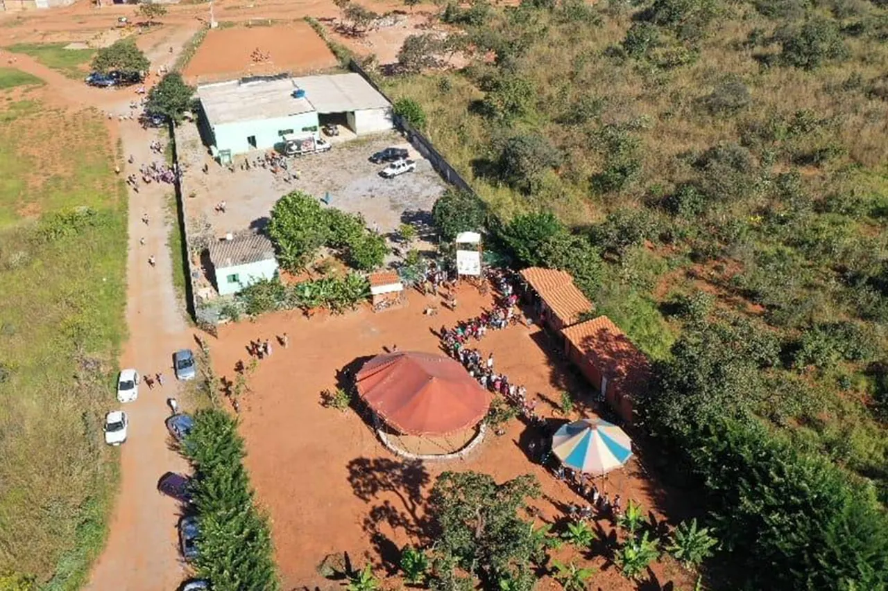 Vista aérea de um terreno de terra vermelha com construções e uma cobertura redonda, cercado de vegetação do Cerrado.