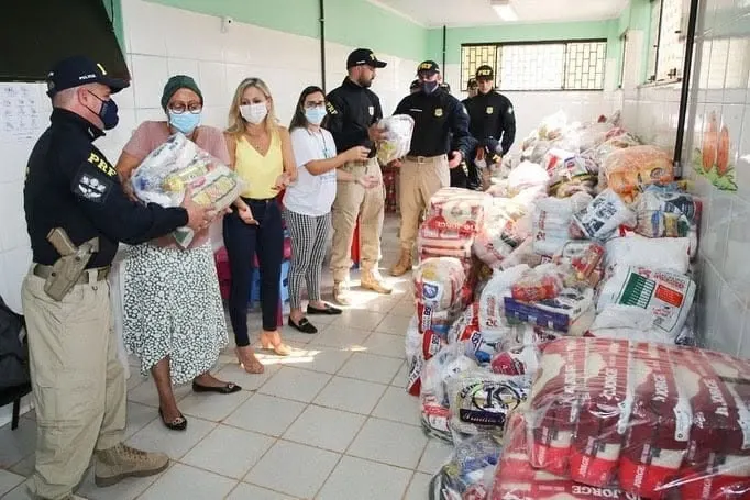 Policiais e voluntárias organizam cestas básicas e mantimentos empilhados em uma sala de azulejos.
