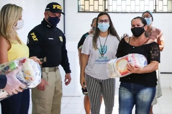 Policial rodoviário federal acompanha a entrega de uma cesta básica a uma moradora, ao lado de voluntárias.