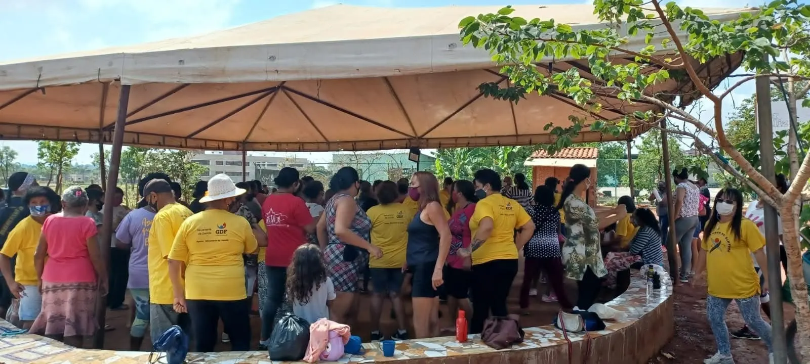 Grupo grande de mulheres, muitas de camiseta amarela, reunido sob uma tenda com banco de mosaico.