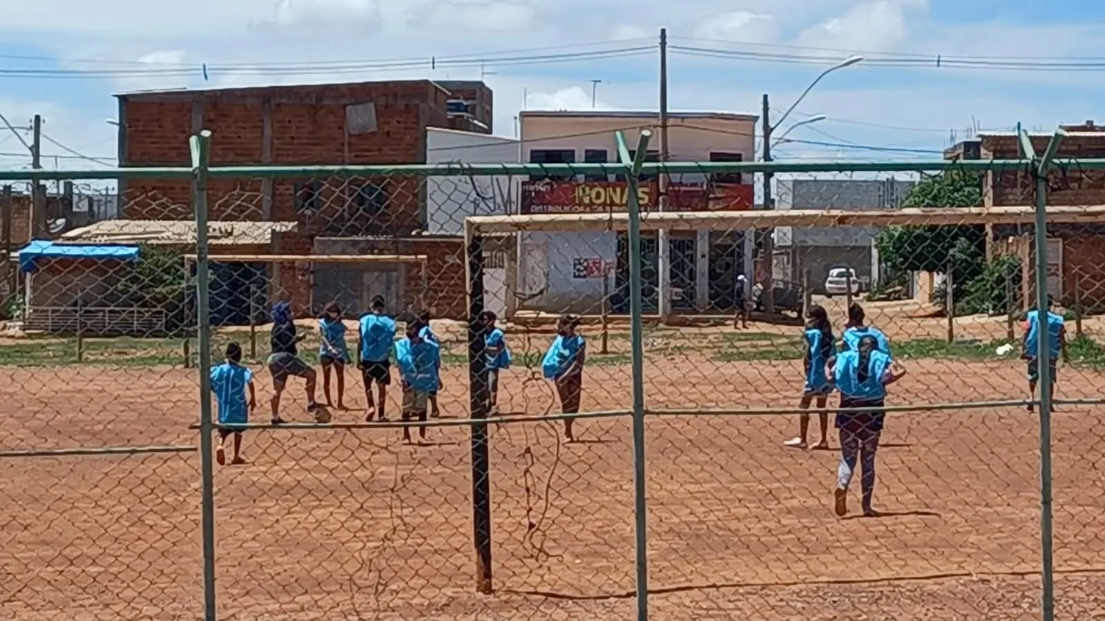Treino de futebol em um campo de terra vermelha, visto de longe através do alambrado, com casas ao fundo.