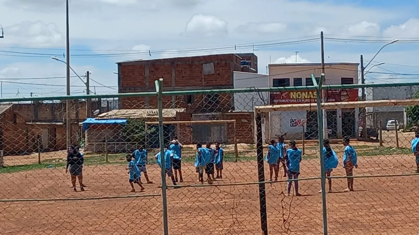 Crianças de colete azul jogam futebol em um campo de terra, vistas de longe através de um alambrado.