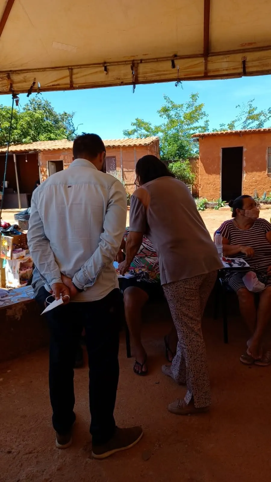 Visitantes, de costas, acompanham o trabalho de artesãs sob a tenda, com casas de barro ao fundo.