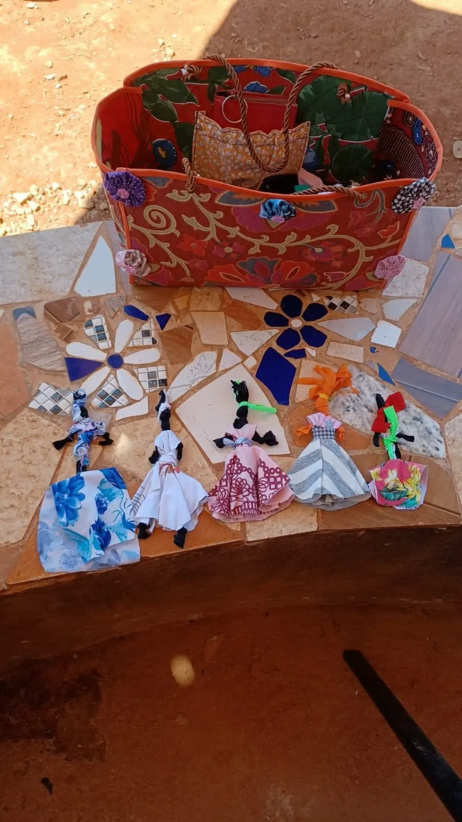 Bonecas de pano feitas à mão e uma bolsa de tecido estampado sobre um banco de mosaico.