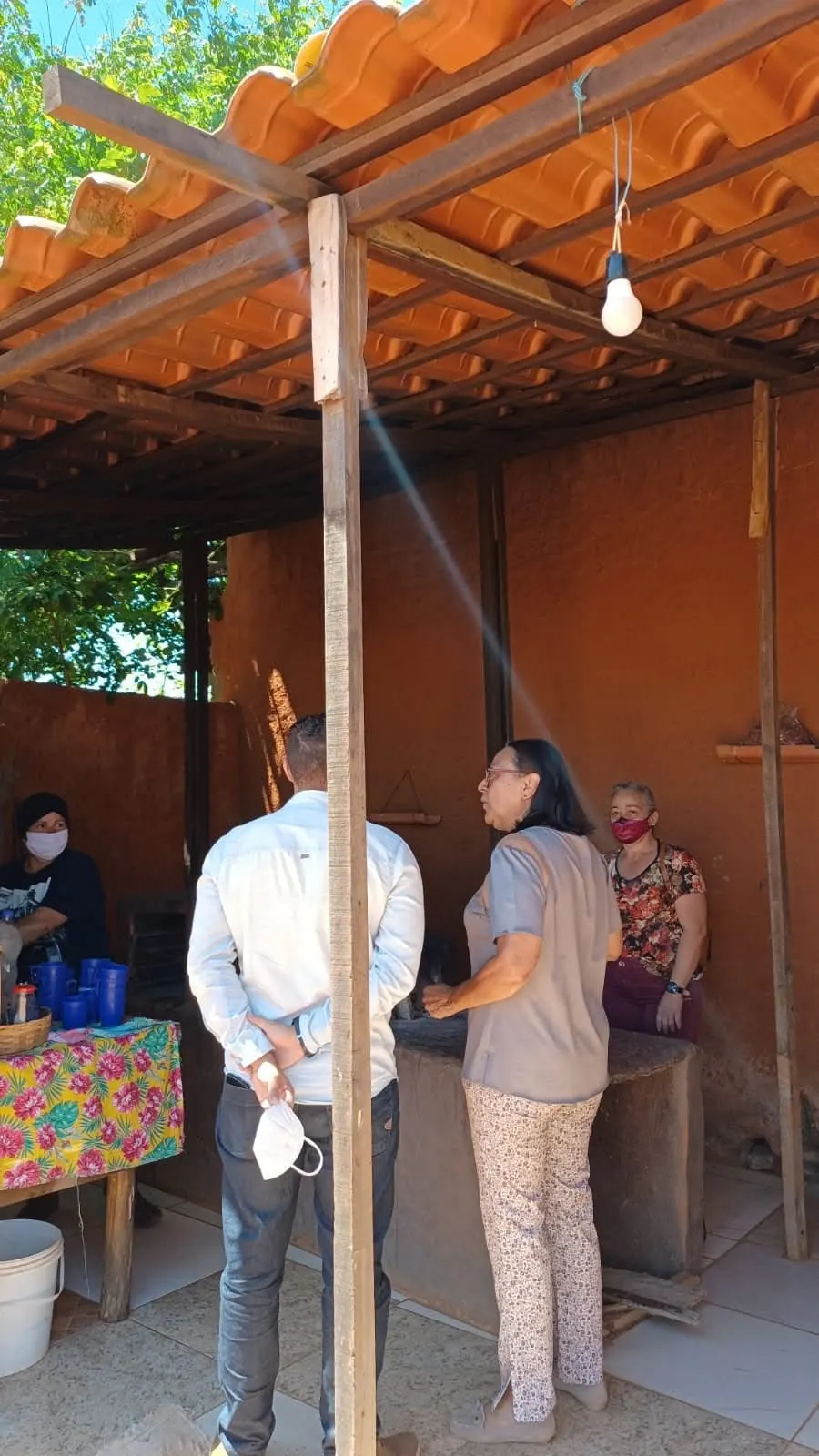 Visitantes conversam com as cozinheiras sob a varanda de telhas de uma cozinha de barro.