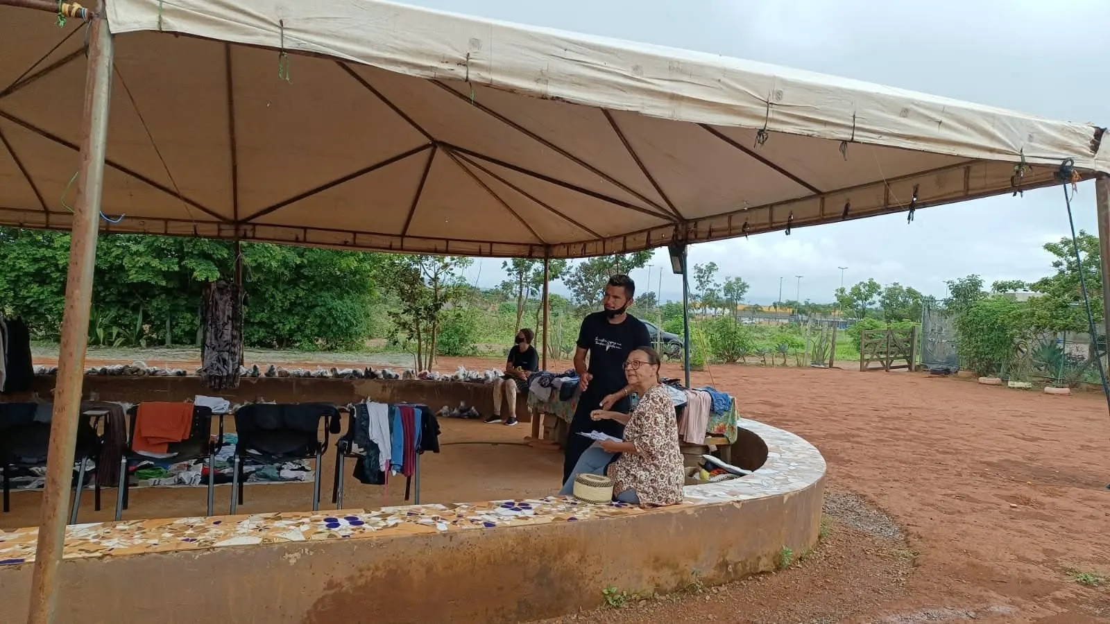 Senhora sentada no banco de mosaico conversa com um jovem sob a tenda do bazar, com terra vermelha ao redor.