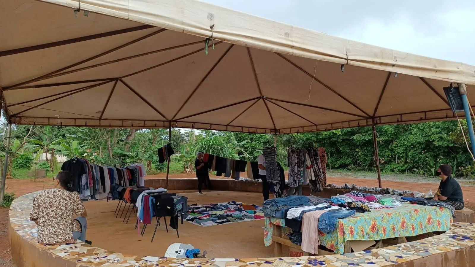 Bazar arrumado sob uma grande tenda com banco de mosaico, com varais de roupas e poucas pessoas.