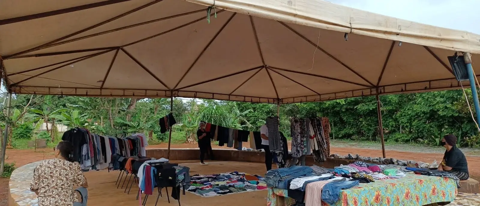 Bazar arrumado sob uma grande tenda com banco de mosaico, com varais de roupas e poucas pessoas.