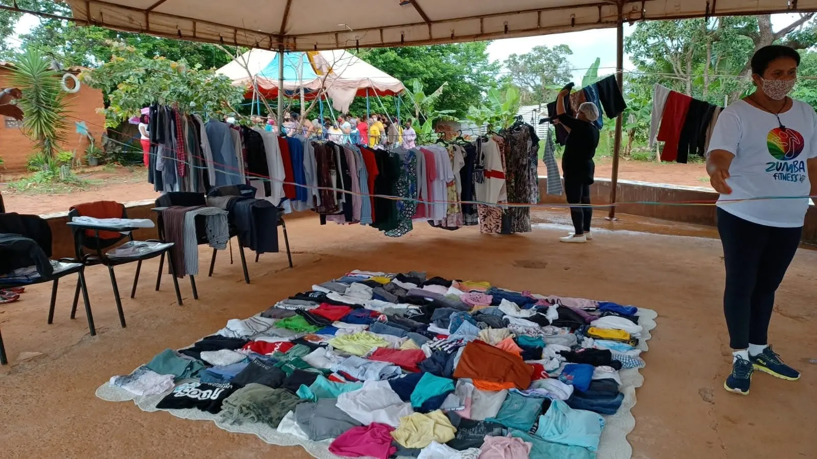 Roupas dobradas sobre uma colcha no chão e penduradas em varais, prontas para o bazar sob a tenda.