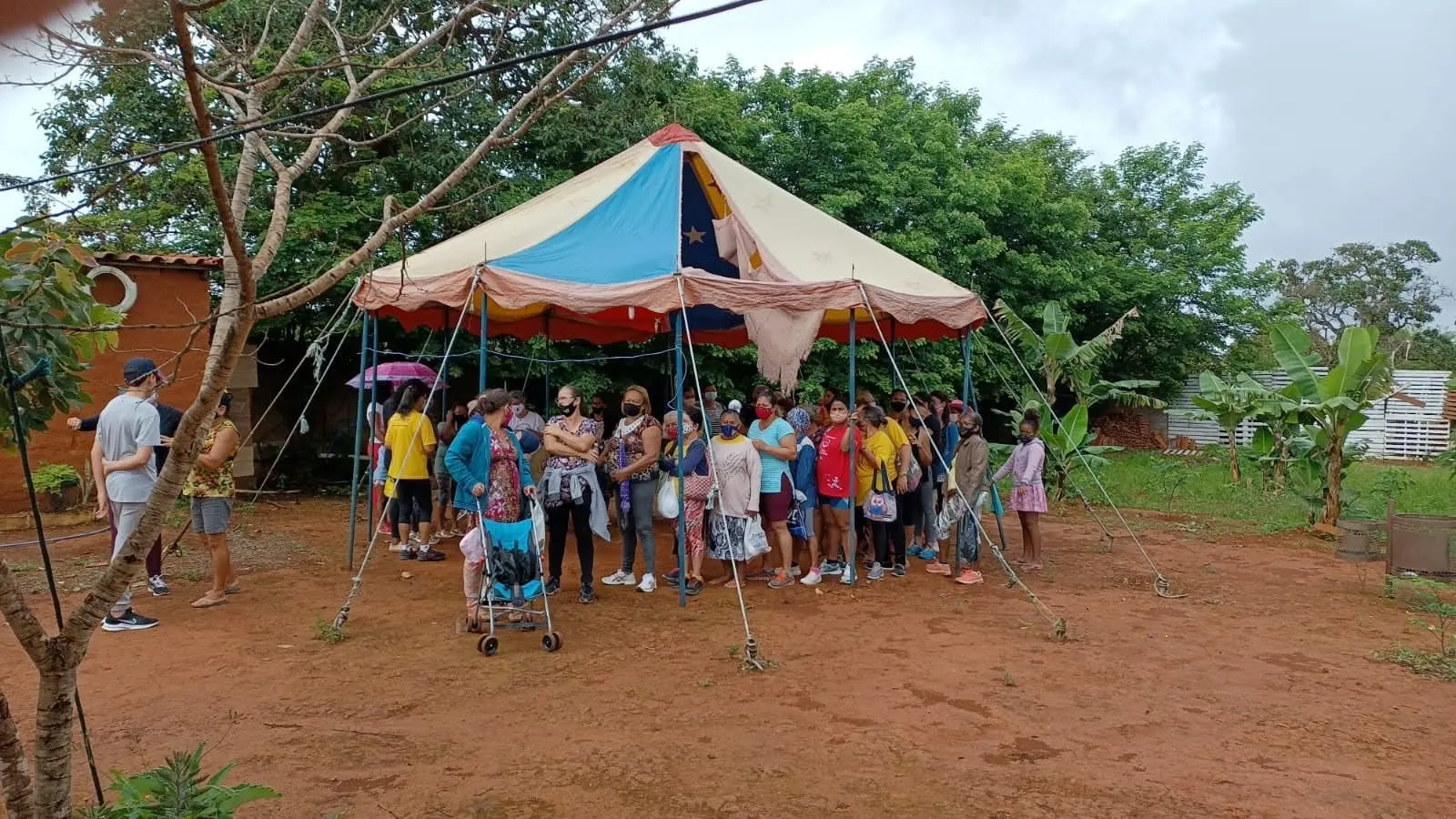 Grupo de moradoras reunido sob uma tenda de circo colorida, cercada de árvores e bananeiras.
