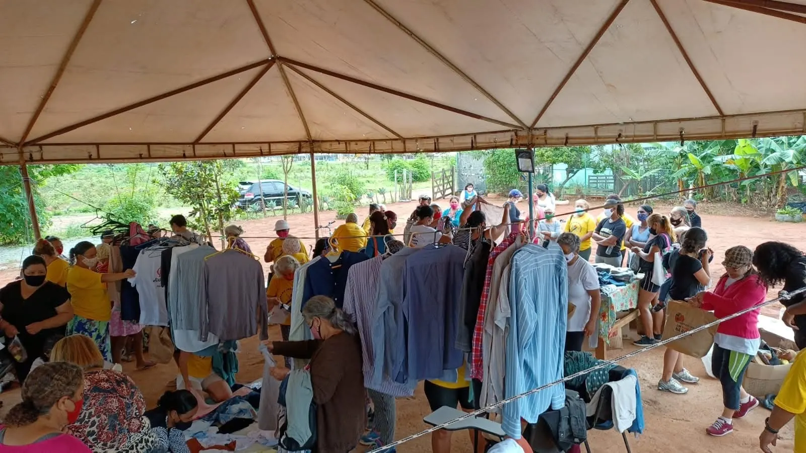 Varais de camisas e mesas de roupas em um bazar movimentado sob uma grande tenda.