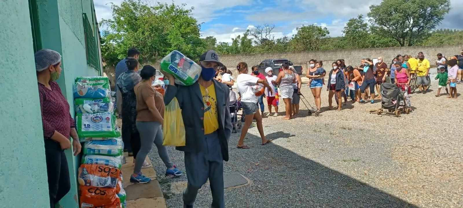 Homem de máscara carrega uma cesta básica no ombro; ao fundo, uma fila de moradores aguarda em um pátio de cascalho.