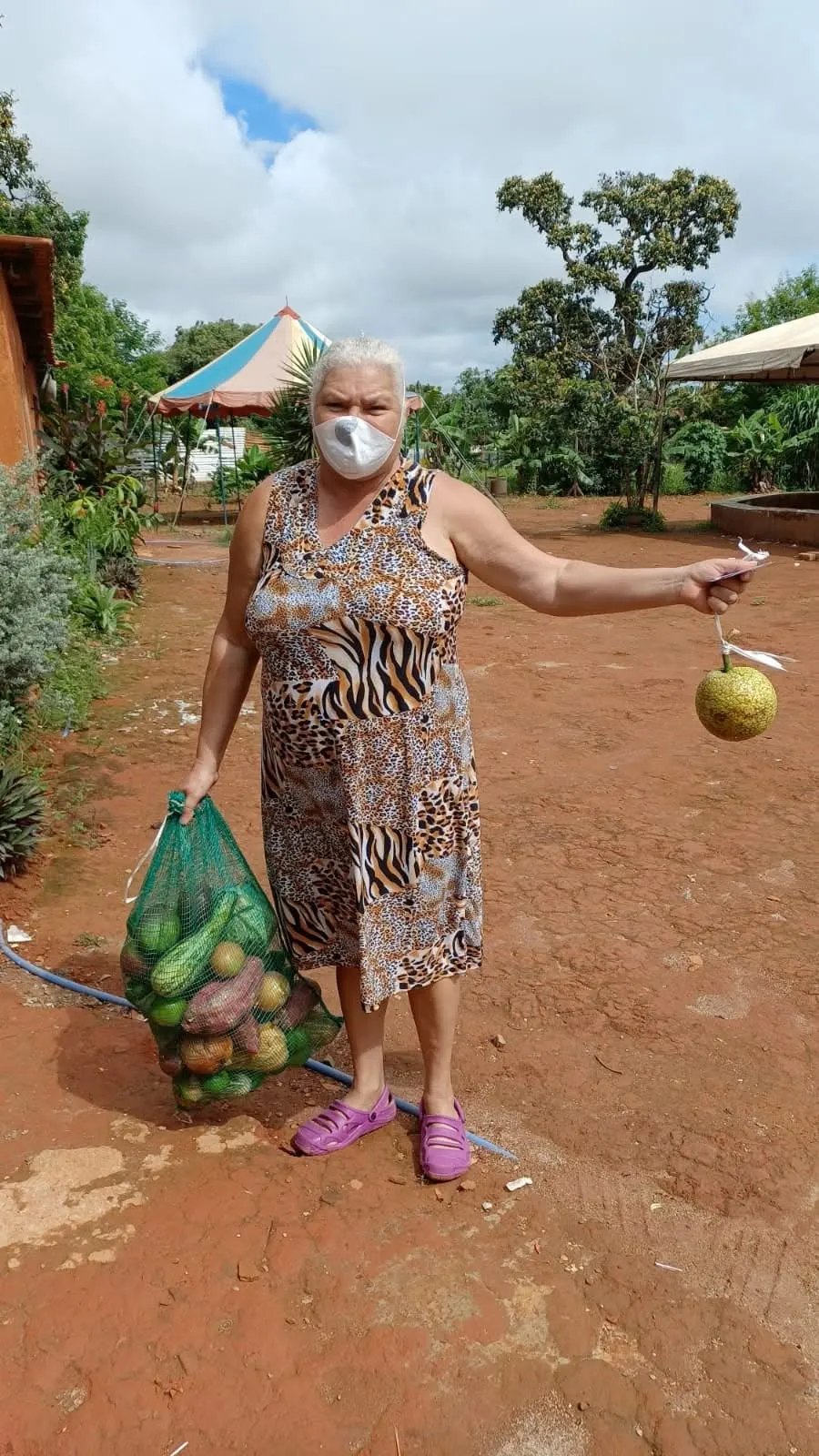 Senhora de vestido estampado segura uma sacola de legumes e uma fruta em um pátio de terra vermelha.
