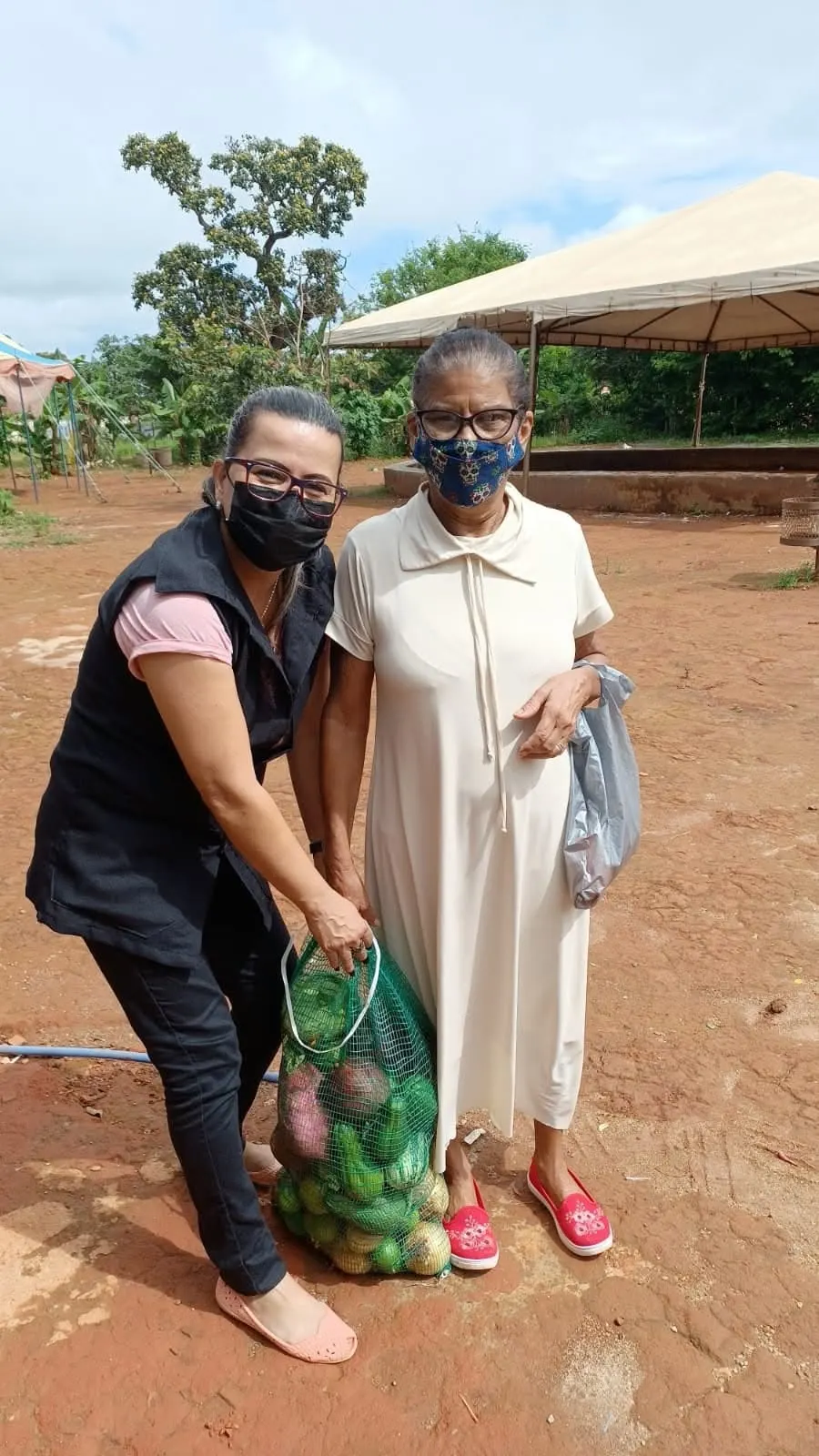 Voluntária e senhora de máscara seguram juntas uma sacola de rede com legumes em um pátio de terra vermelha.