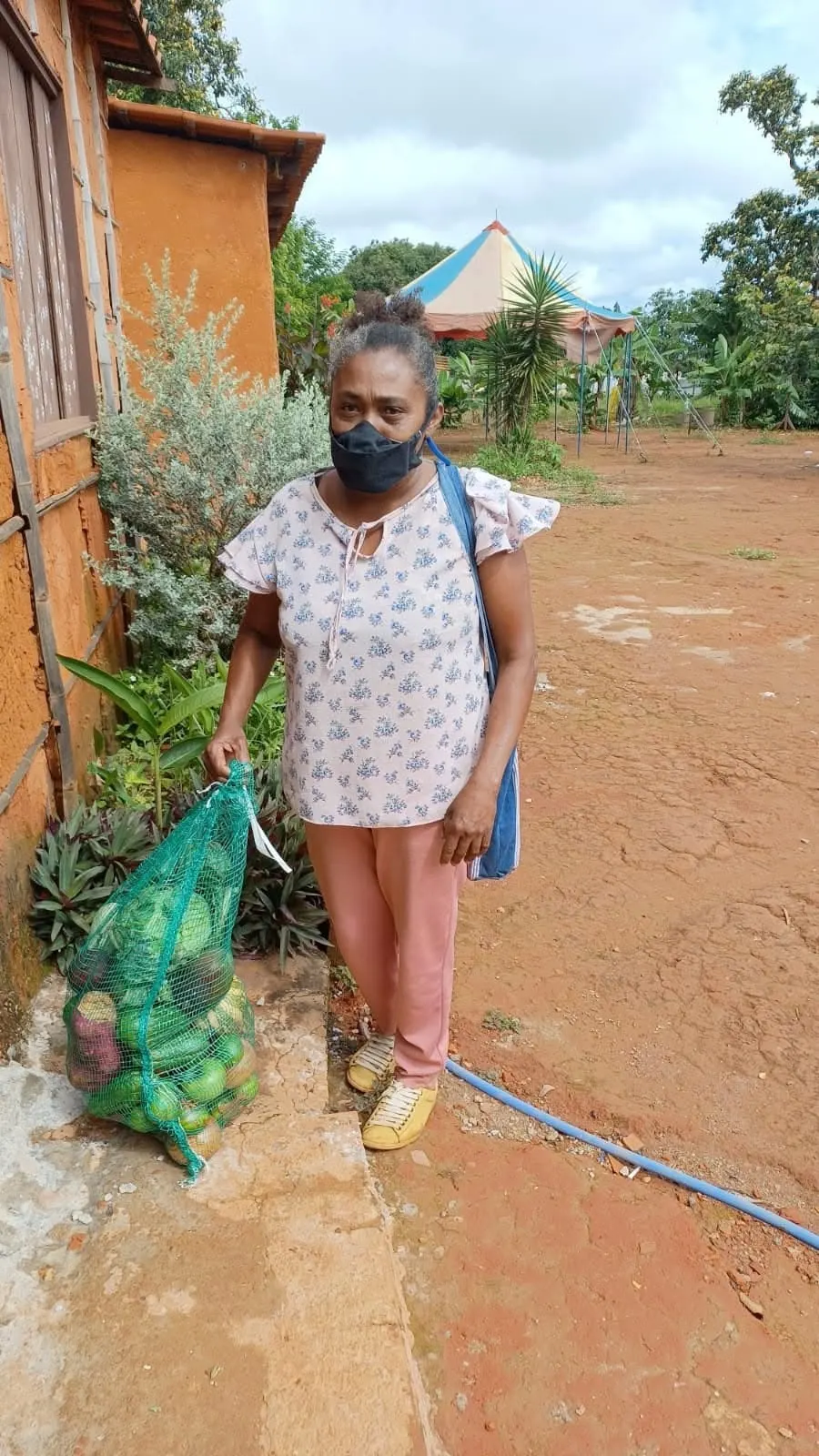 Senhora de máscara ao lado de uma sacola de rede cheia de legumes, junto a uma casa de barro.