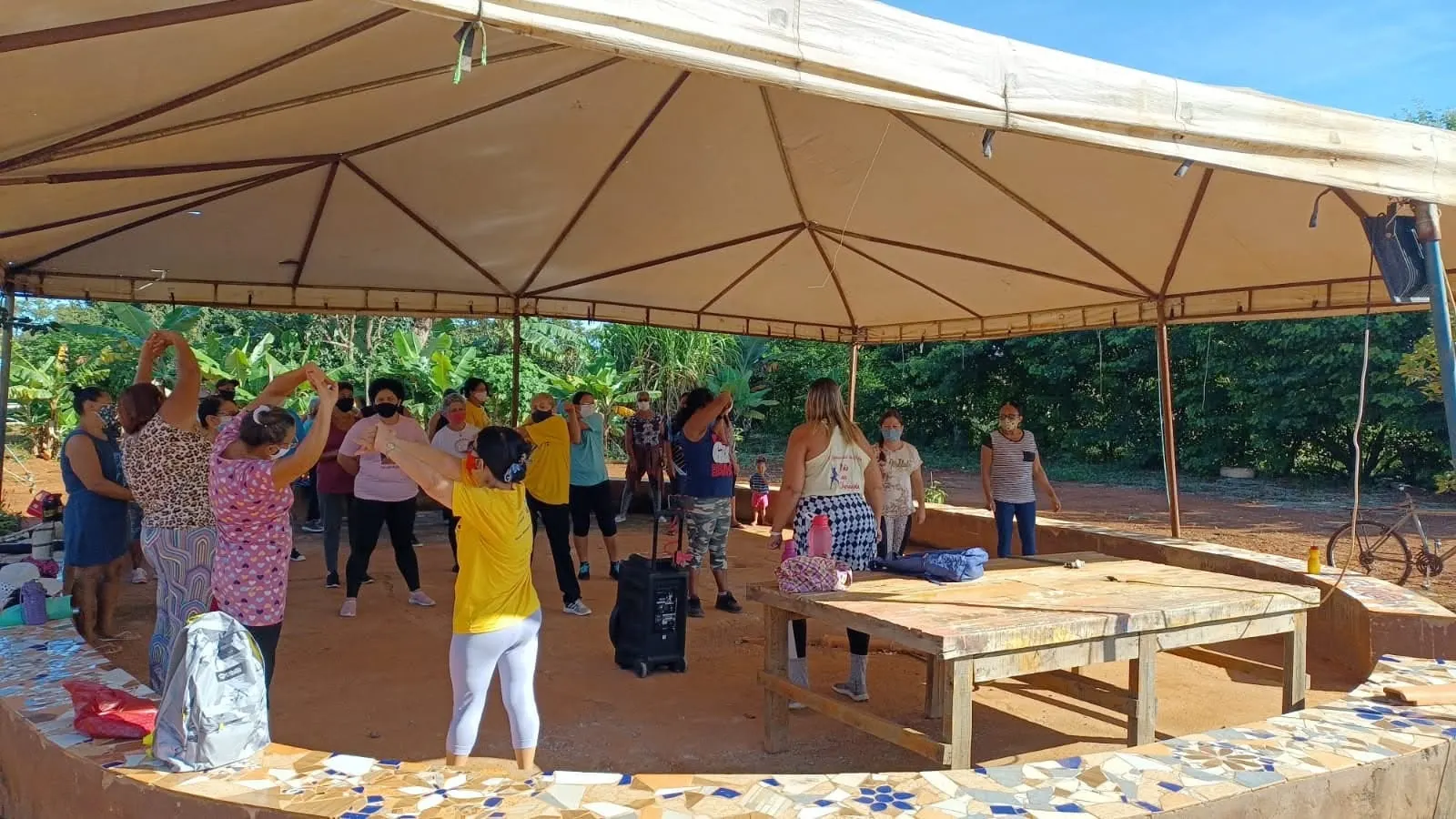 Mulheres alongam os braços em uma aula de zumba sob uma grande tenda com banco de mosaico.