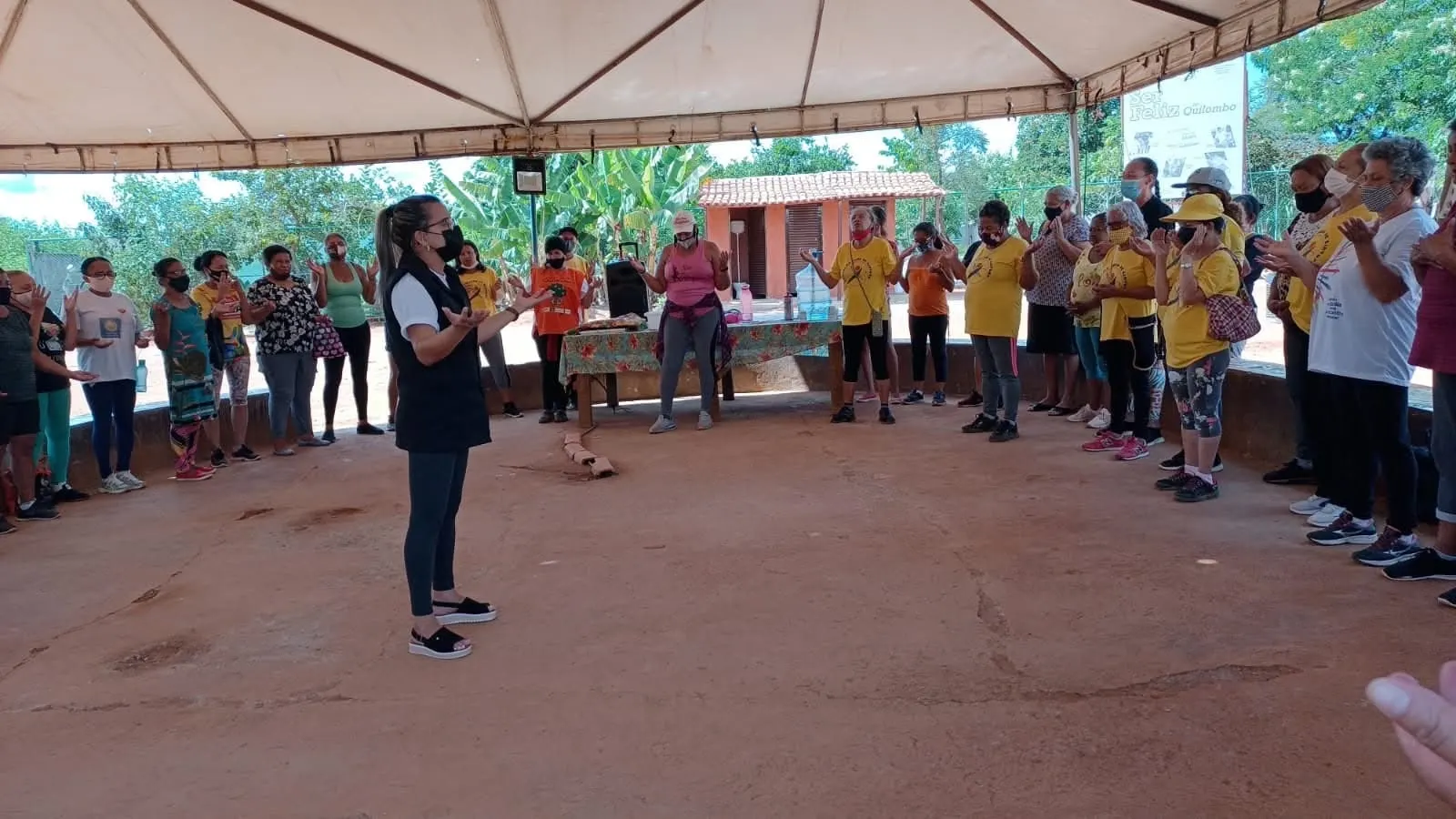 Mulheres em roda sob uma grande tenda, de mãos abertas, acompanham uma orientadora.