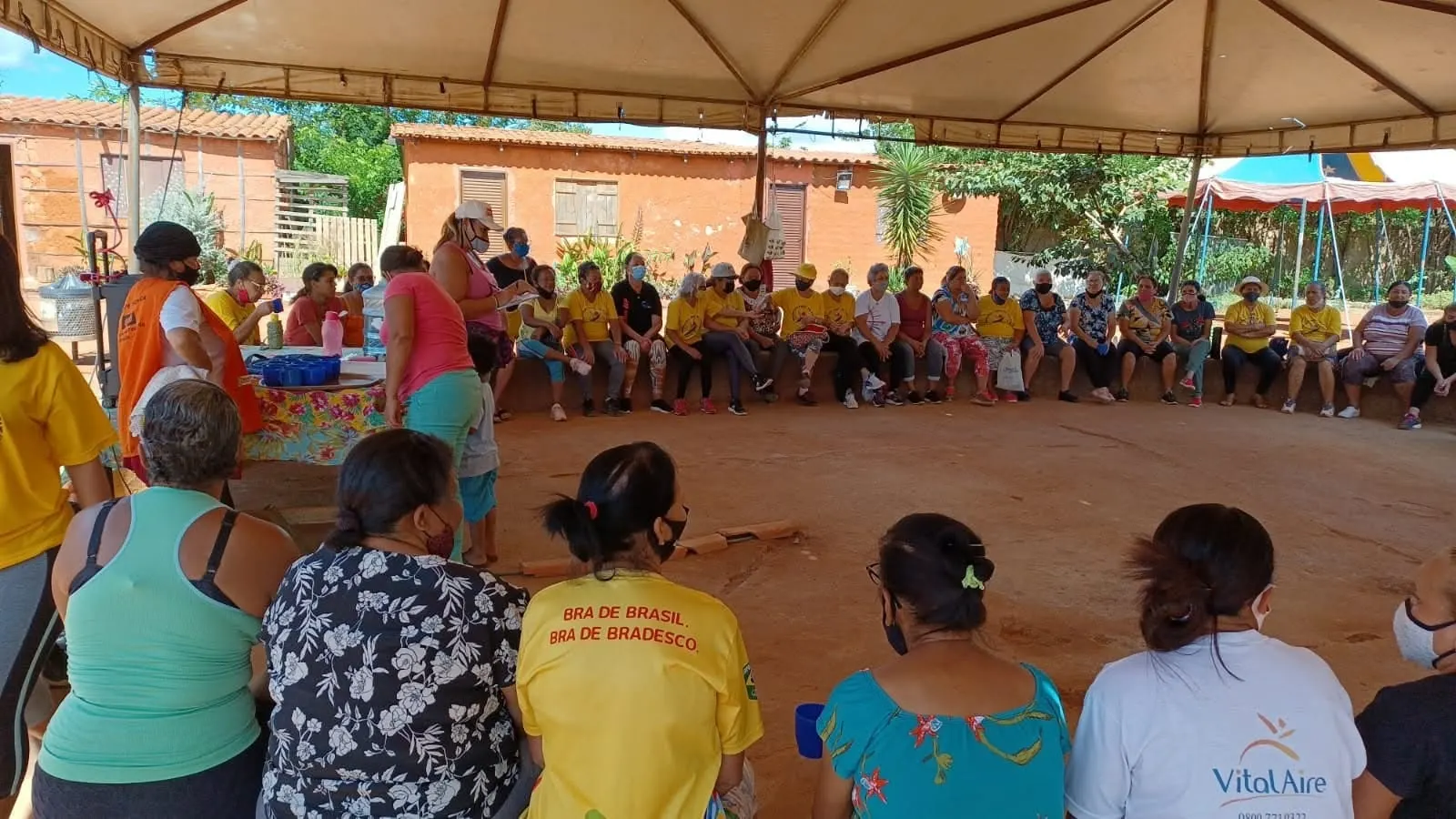 Roda de conversa com idosas sentadas em volta de um espaço de terra sob uma grande tenda, vistas de costas.