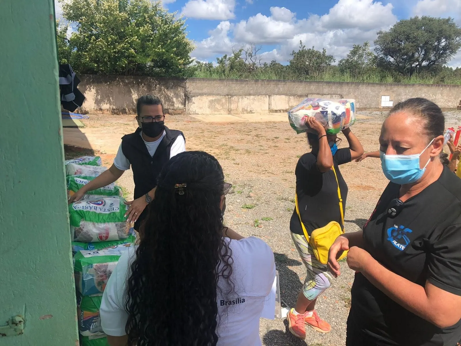 Voluntária de máscara organiza cestas básicas junto à parede verde enquanto uma senhora leva a sua cesta na cabeça.
