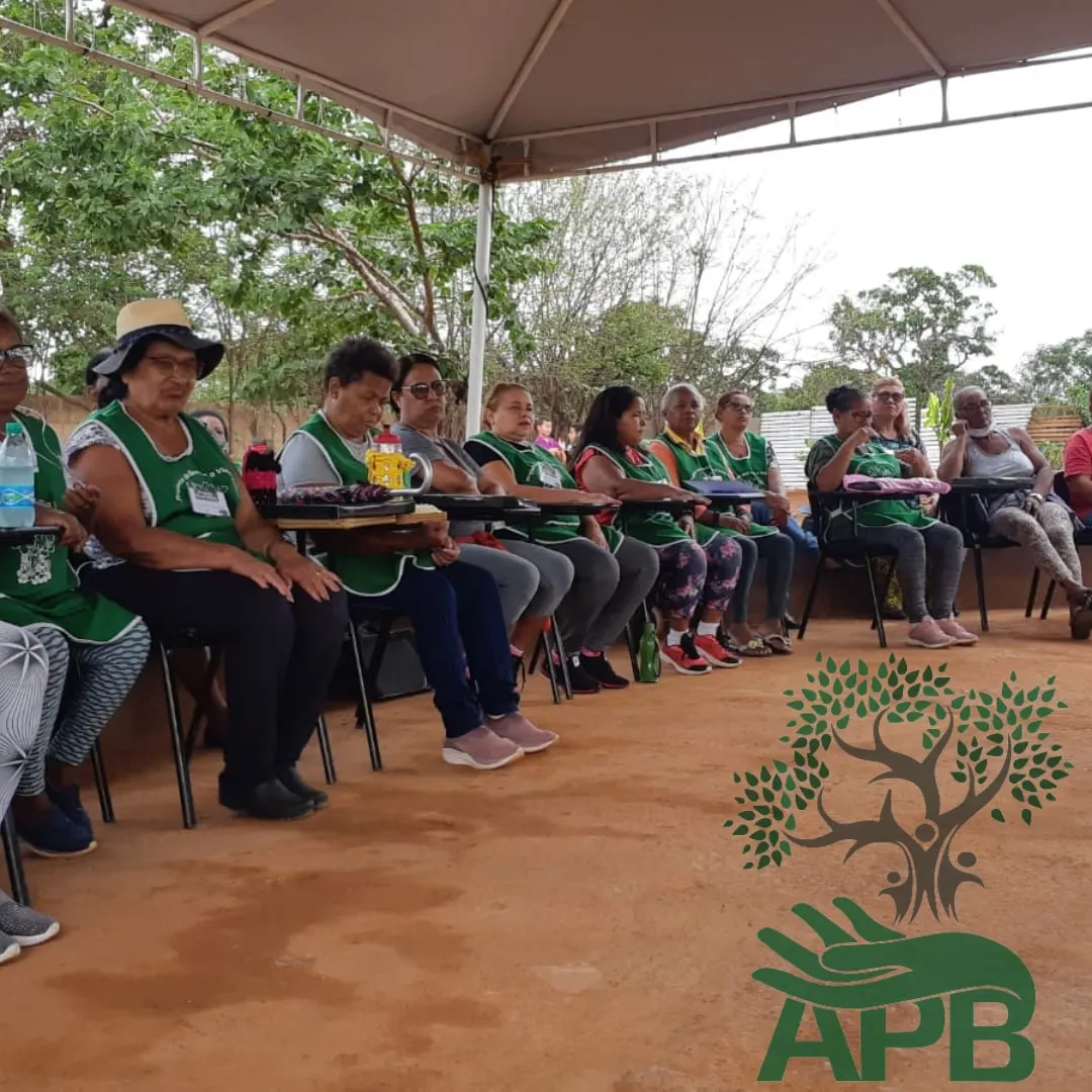 Idosas de avental verde sentadas em fila sob uma tenda ao ar livre.