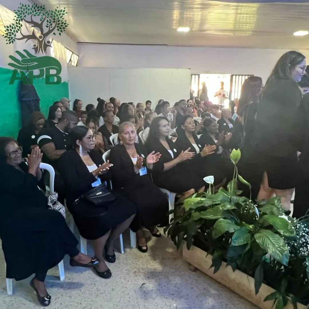 Plateia de mulheres vestidas de preto aplaudindo, sentadas em cadeiras brancas durante uma cerimônia.