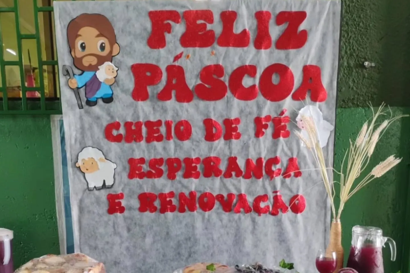 Mesa de Páscoa com pães e frutas diante de um painel com a frase "Feliz Páscoa, cheio de fé, esperança e renovação".
