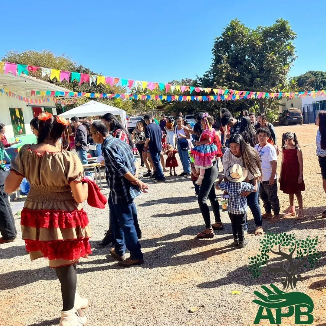 Festa junina ao ar livre com bandeirinhas coloridas, pessoas em trajes típicos e crianças ao longe.