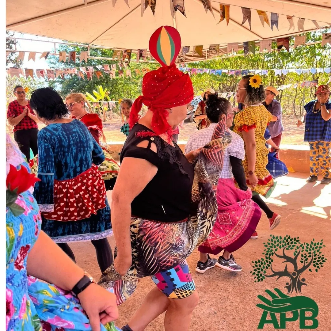 Adultos dançam em trajes de festa junina sob a tenda, com um chapéu vermelho em destaque.