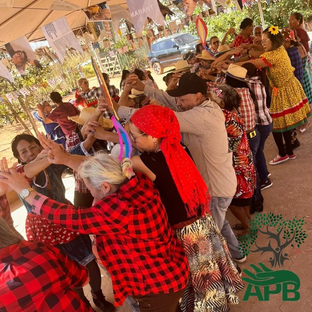 Adultos em trajes de festa junina dançam em fila sob uma cobertura.