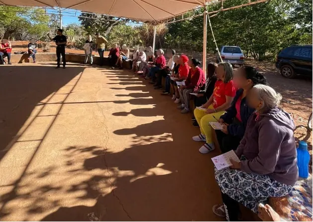 Participantes sentados em fila à sombra ouvindo um palestrante em pé ao fundo.
