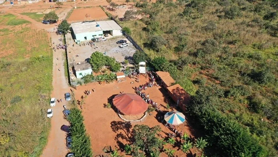 Vista aérea da sede da APB em Itapoã, com construções, uma quadra de terra e uma cobertura redonda cercadas de vegetação.